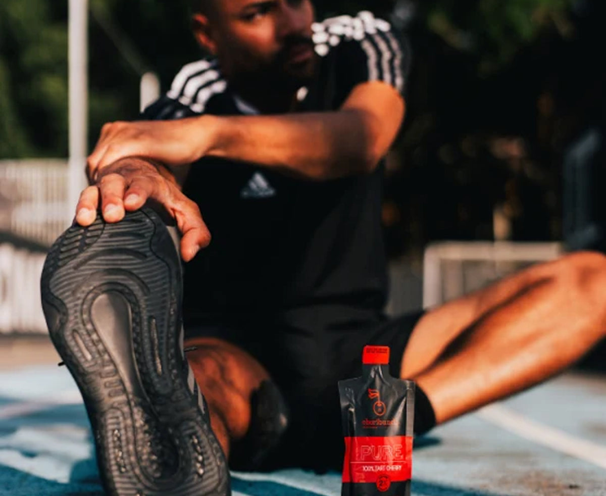 A man sitting on a track wearing a black Adidas athletic outfit and stretching, with a small red-and-black Cheribundi hydration pouch placed in the foreground on the track.