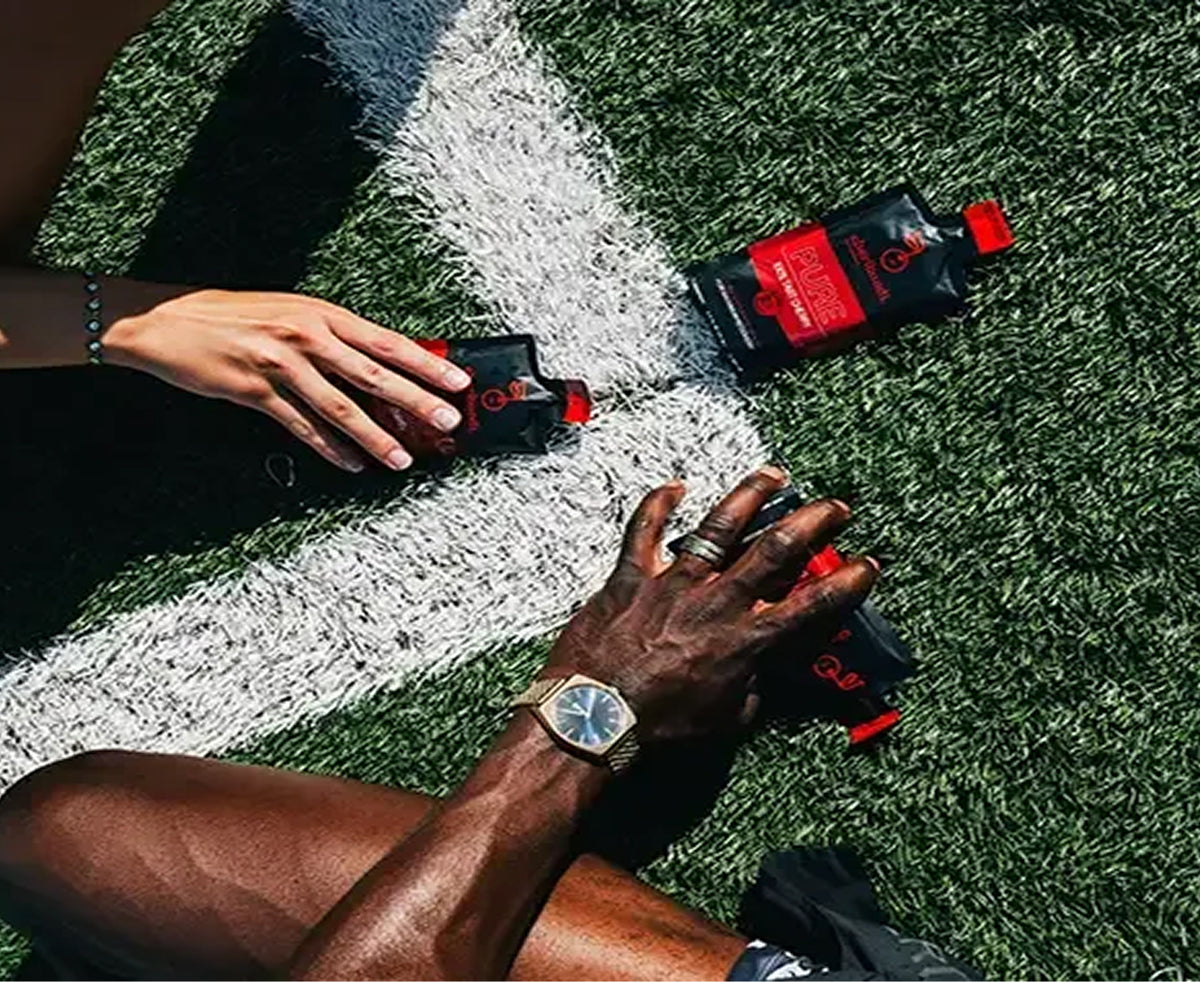 Two black and red sports drink pouches are placed on green turf near a white yard line, with two human hands reaching toward them. One hand wears a gold watch and ring.